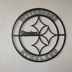 Op de afbeelding: Een cirkelvormig wandbord van zwart metaal met het logo van de Pittsburgh Steelers. Het ontwerp bevat de tekst Pittsburgh aan de bovenkant, Steelers aan de onderkant en het woord Steelers in het midden, omringd door de iconische vierpuntige stervormen.