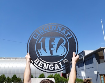 Pode incluir: Uma pessoa segura um sinal de metal preto e azul com as palavras "Cincinnati Bengals" e um "B" estilizado no centro.