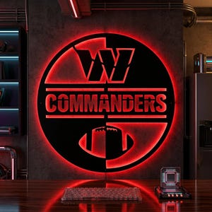 May include: A circular black metal wall sign featuring the Washington Commanders logo with a stylised W, the text COMMANDERS, and a football graphic, all illuminated from behind by a vibrant red LED light glow against a dark textured wall.