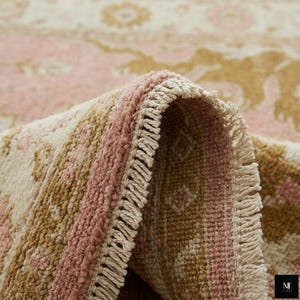 May include: Close-up of a rolled-up area rug with a floral pattern. The rug features a color palette of pink, beige, and tan. The edges of the rug have a fringe detail. The rug is made of a soft, woven material.