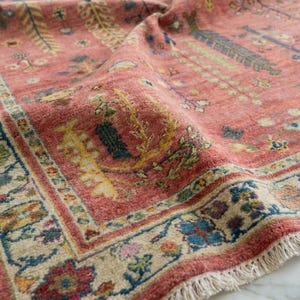 Puede incluir: Primer plano de una alfombra vintage con un fondo rojo descolorido y intrincados diseños florales en amarillo, azul y verde. La alfombra tiene un borde con flecos y un borde detallado.