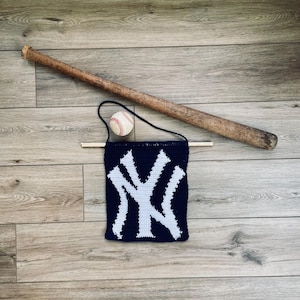 Könnte beinhalten: Ein gehäkeltes Wandbehang mit dem Logo der New York Yankees, einem Baseball und einem Holzschläger auf einem Holzboden.