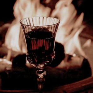 Może przedstawiać: Kieliszek wypełniony ciemnoczerwonym płynem, prawdopodobnie winem, znajduje się na pierwszym planie. Kieliszek ma szczegółowy, ozdobny wzór. Tło jest rozmyte, z nutami ognia lub płomieni, tworząc dramatyczny, ciepły obraz.