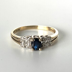 May include: A gold ring with a central oval, dark blue gemstone, and two clusters of smaller, clear gemstones. The polished gold band has a split shank design. The ring is set against a white background. The ring is likely made of gold.