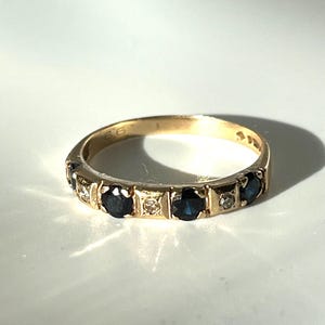 May include: A gold ring featuring alternating dark blue and clear gemstones. The ring, with a classic design, is set against a white backdrop. The number "66" is engraved on the ring.