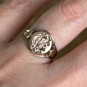 May include: A gold signet ring featuring a circular face with an engraved monogram of the letters M and A intertwined. The band is smooth and polished, showcasing a warm metallic tone throughout the entire piece of jewellery.
