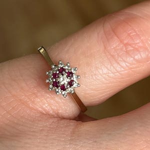 May include: A gold ring featuring a central cluster of small, round, deep red gemstones, encircled by smaller, clear stones. The ring has a thin, gold band, and the gemstone setting is silver-coloured.