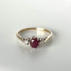 May include: A gold ring with an oval ruby centre stone and two smaller clear gemstones either side. The ring has a slender band and a classic design. The ring is suitable for everyday wear or special occasions.