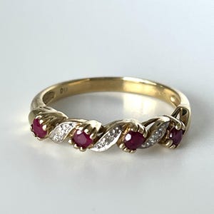 May include: A gold band ring featuring a twisted design with four round-cut red rubies alternating with small white diamonds set in silver-toned metal. The interior of the band is engraved with the letters DIA, indicating a gold and diamond construction.
