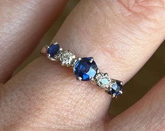 May include: A five-stone ring featuring a central round-cut blue sapphire flanked by two smaller round-cut diamonds and two outer round-cut blue sapphires, all set in a polished metal band with prong settings arranged in a linear design.