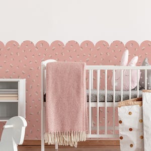 Op de afbeelding: Een kinderkamer met een witte wieg, een roze deken en decoratieve kussens. De kamer heeft roze bloemenbehang en twee papieren opbergzakken, een met gouden stippen en de andere met gesloten ogen.