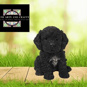 May include: A black puppy figurine with a fluffy coat and a small white patch on its chest. The puppy is sitting on a wooden surface with green grass in the foreground and a blurred green background. The image includes a logo that says "North Arts and Crafts".