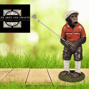 May include: A whimsical figurine of a chimpanzee golfer, wearing a red polo shirt, black shorts, and a white cap. The chimp is holding a golf club, standing on a patch of green grass. The background is a blurred green.