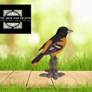 May include: A detailed figurine of an oriole bird perched on a tree branch. The bird features black, orange, and white plumage. The background includes a logo that reads "North Arts and Crafts" and a blurred green and brown backdrop.