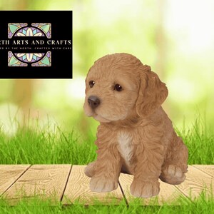 May include: A light brown puppy figurine with a fluffy coat, sitting on a wooden surface. The puppy has a white patch on its chest and is looking to the left. The background is blurred with green grass and a logo.