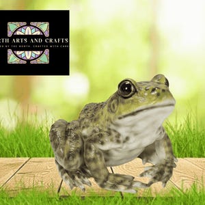 May include: A detailed frog ornament with a textured, green and brown finish. The frog is positioned on a wooden surface with green grass in the foreground and a blurred green background. The image includes the text "NORTH ARTS AND CRAFTS".