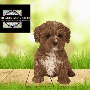 May include: A brown puppy figurine with a white patch on its chest sits on a wooden surface. The puppy has curly fur and dark eyes. The background is a blurred green and white, with a logo that reads "NORTH ARTS AND CRAFTS".