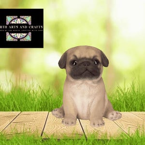 May include: A tan pug puppy figurine with a black muzzle and dark eyes, sitting in a forward-facing position. A black square logo in the upper left corner displays the text NORTH ARTS AND CRAFTS INSPIRED BY THE NORTH, CRAFTED WITH CARE.