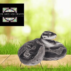 May include: A coiled snake with a black and grey pattern rests on a wooden surface with green grass in the foreground. The snake's head is raised, and the background is a blurred green. A logo for North Arts and Crafts is in the upper left corner.