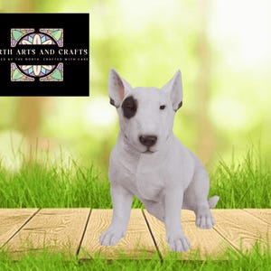 May include: A white Bull Terrier ornament with a black patch over one eye is positioned on a wooden surface with green grass in the foreground. The background is a blurred green and white. A black square in the upper left corner displays the text "NORTH ARTS AND CRAFTS".