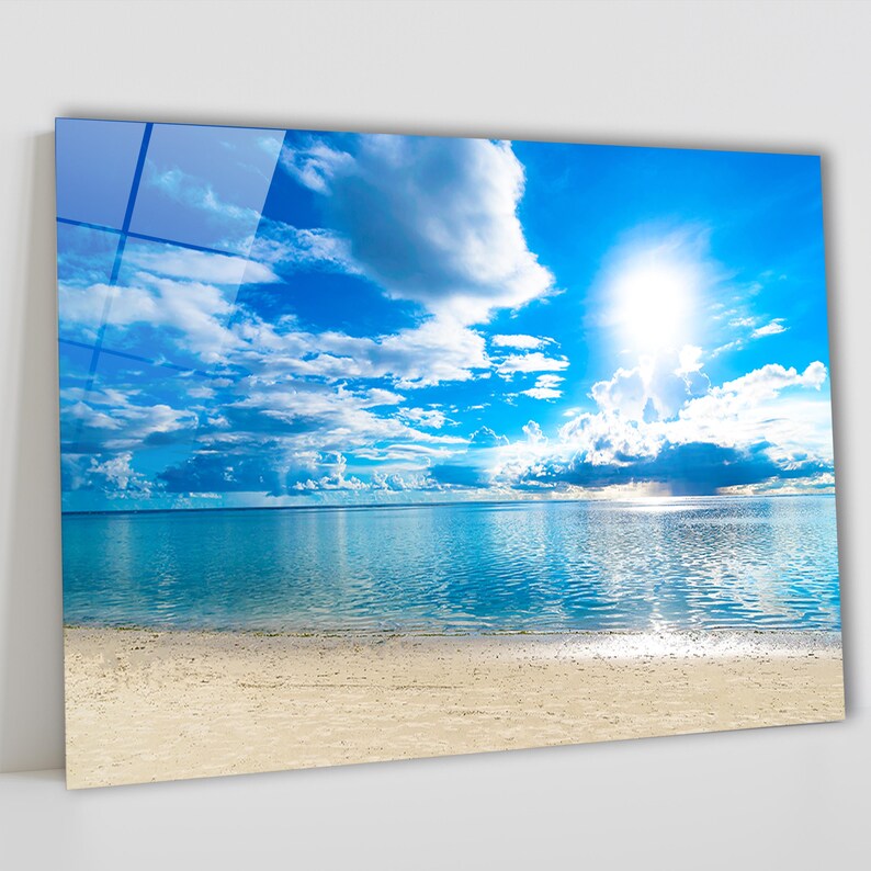 May include: A vibrant image of a sunny beach scene. The sky is a brilliant blue, dotted with fluffy white clouds. The ocean is a calm turquoise, meeting a sandy beach. The sun shines brightly, reflecting on the water, creating a serene and picturesque landscape.