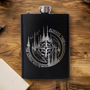 May include: Black stainless steel flask with a silver compass and mountain design. The text reads "ALWAYS TAKE THE SCENIC ROUTE." The flask has a hinged cap and is likely used for carrying spirits or other beverages.