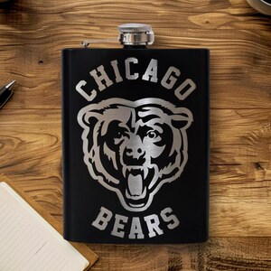 May include: A black stainless steel flask with a silver Chicago Bears logo and text. The flask has a hinged cap and is set against a wooden background. The flask is rectangular and appears to be empty.