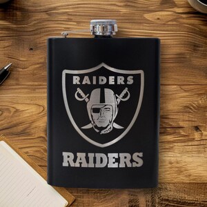 May include: Black stainless steel flask with a silver Raiders logo and text. The logo features a helmeted face with crossed swords and the word "RAIDERS" above and below the emblem. The flask has a silver cap.