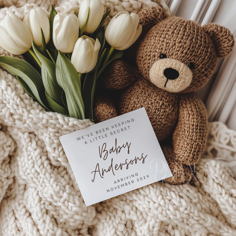 May include: A knitted brown teddy bear rests on a cream-colored blanket next to a bouquet of white tulips. A white card with the text "We've been keeping a little secret, Baby Andersons, Arriving November 202X" is in the foreground.