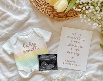May include: A white onesie with a rainbow ombre design and the text "baby smith" in brown script. A black and white ultrasound image is next to the onesie. A white card with the text "We're expecting a new addition to our family arriving October 2025" is on a white surface with a bouquet of white and yellow flowers.