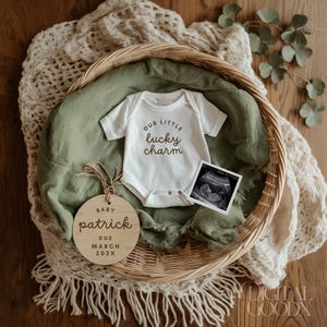 May include: A white baby onesie with the text "Our little lucky charm" in brown lettering. A wooden circle with the text "Baby Patrick Due March 202X" is next to the onesie. A black and white ultrasound image is also in the photo.