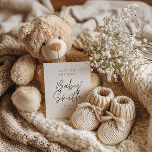 May include: A beige teddy bear, baby booties, and a card that reads "Baby on the way" with the name "Baby Smith" and the due date "01/2025". The items are arranged on a cream-coloured knitted blanket with white flowers.