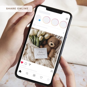 May include: A smartphone displays a baby announcement with a teddy bear, white tulips, and a knitted blanket. The announcement reads "Baby Andersons" and "Arriving November 2023". The phone screen also shows social media icons.