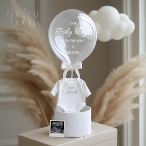 Puede incluir: Un body blanco para bebé con el texto "Baby Smith" está unido a un globo transparente con el texto "Our Baby Boy is on the way! 08.2025" y una nube blanca. El globo está sentado en una caja blanca con una pequeña imagen de ultrasonido.