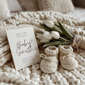 May include: A cream-colored announcement card with the text "Baby on the Way" and "Baby Smith Arriving Dec 2025" sits next to a bouquet of white tulips and a pair of knitted baby booties on a cream-colored blanket.