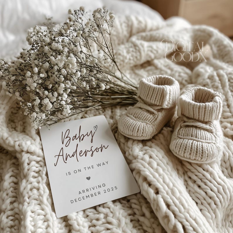 Peut inclure: Une paire de chaussons de b&eacute;b&eacute; en tricot cr&egrave;me &agrave; c&ocirc;t&eacute; d'une carte indiquant "Baby Anderson is on the way, arriving December 2025." Les articles sont sur une couverture en tricot cr&egrave;me, avec une branche de fleurs blanches.