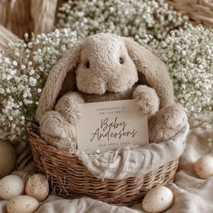 May include: A soft, beige bunny toy in a wicker basket, holding a card with the text "Somebunny is on the way" and "Baby Andersons, Due April 202X." The basket is lined with fabric and surrounded by speckled eggs and white flowers.