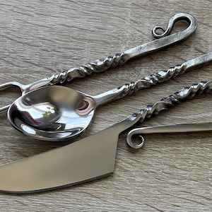 May include: A set of handcrafted metal serving utensils, including a fork, spoon, and knife. The utensils have twisted handles and decorative curled ends. The polished metal reflects light, and the set is arranged on a wooden surface.