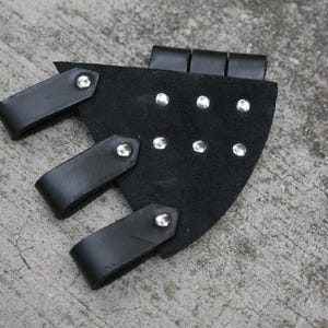 May include: Black leather bracer with three buckle straps and silver rivets. The bracer is made of a thick, durable material and is designed to protect the forearm. The bracer is a piece of armor, often used in cosplay or historical reenactments.