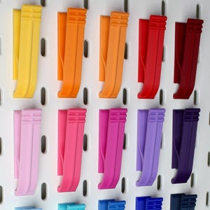 Könnte beinhalten: Eine Sammlung farbenfroher Lochwandhaken in verschiedenen Farbtönen, darunter Gelb, Orange, Rot, Rosa, Lila, Blau, Grün und Schwarz. Die Haken sind auf einer weißen Lochwand angeordnet und zeigen ihre vielfältige Farbpalette.