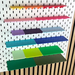 May include: A white rectangular pegboard featuring a grid of vertical slots. The board is equipped with multiple small, flat shelves in a spectrum of colors including yellow, orange, red, pink, purple, light blue, dark blue, teal, lime, and green.