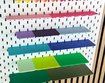 May include: A white rectangular pegboard featuring a grid of vertical slots. The board is equipped with multiple small, flat shelves in a spectrum of colors including yellow, orange, red, pink, purple, light blue, dark blue, teal, lime, and green.