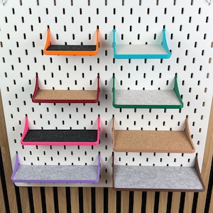May include: A white rectangular pegboard featuring a grid of vertical slots. Eight small shelves in various colors, including orange, teal, red, green, pink, brown, and purple, are attached to the board, each with a flat surface insert.