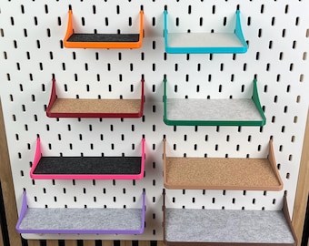 May include: A white rectangular pegboard featuring a grid of vertical slots. Eight small shelves in various colors, including orange, teal, red, green, pink, brown, and purple, are attached to the board, each with a flat surface insert.