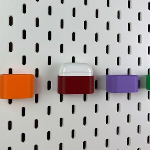 May include: A white pegboard with vertical slots features several small, colorful plastic storage containers attached to it. One container is bright orange, one is deep red holding white wireless earbuds, and one is purple, all mounted securely.