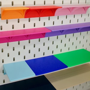 Puede incluir: Un conjunto de estantes de plástico de colores con bordes redondeados. Los estantes están montados en una pared de tablero de clavijas blanco. Los estantes están en los siguientes colores: naranja, rojo, rosa, morado, azul claro, azul oscuro, verde y beige.