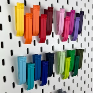 May include: A collection of multi-colored plastic clips attached to a white pegboard with vertical slots. The clips are arranged in rows and feature a variety of vibrant hues including yellow, orange, red, pink, purple, blue, and green.