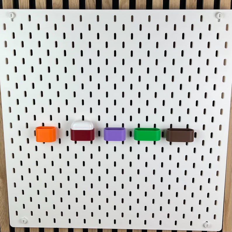 May include: A white rectangular pegboard with vertical slots features five small, colorful storage holders attached in a horizontal row. The holders are orange, a white-topped dark red, purple, green, and brown, providing organized storage.