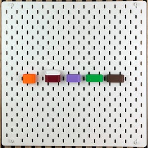 May include: A white rectangular pegboard with a grid of vertical slots features five small, colorful storage containers attached in a horizontal row. The containers are orange, maroon with a white lid, purple, bright green, and dark brown.