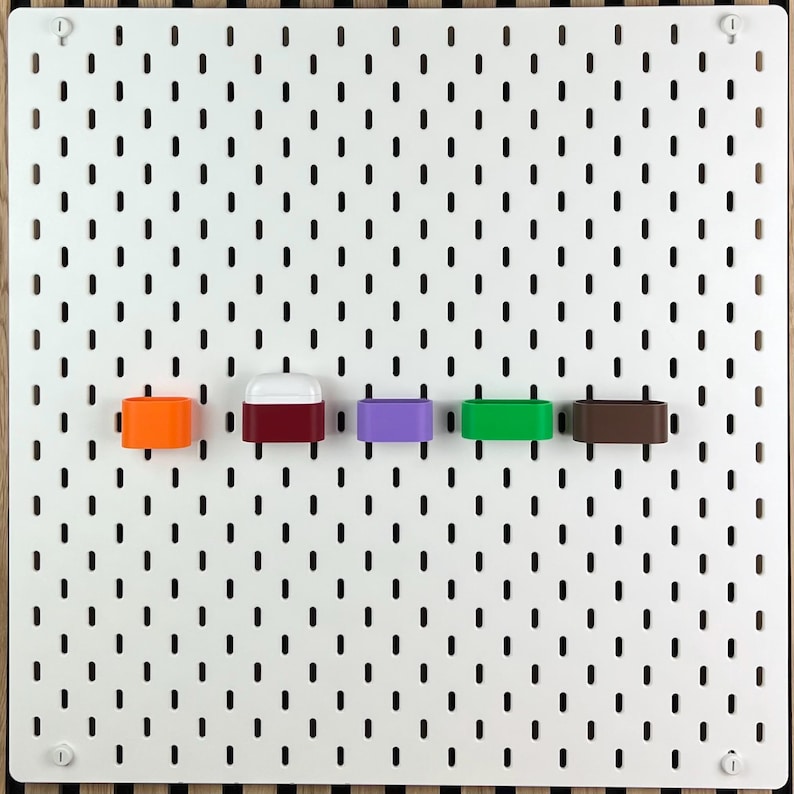 May include: A white rectangular pegboard with a grid of vertical slots features five small, colorful storage containers attached in a horizontal row. The containers are orange, maroon with a white lid, purple, bright green, and dark brown.